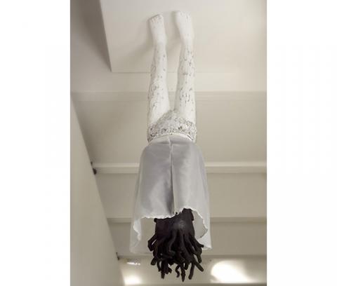 female figure, bottom half white, top half painted black, spiky hair, hands folded over chest, white painted metal underpants and white cloth skirt; figure is suspended from ceiling with skirt covering upper half body