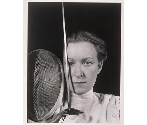 bust portrait of woman in fencing uniform, holding sword vertical over right eye and mask beside that