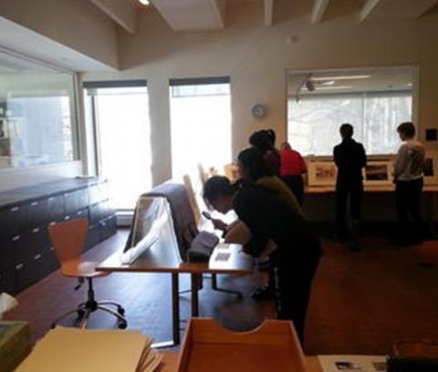 tables set up around a room with artworks propped up on them. students look closely at the artworks using magnifying glasses