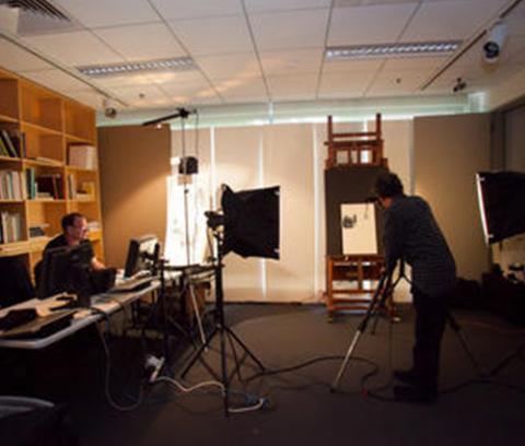man sitting at a desk with a bookshelf behind him. to the right are two large box lights projecting onto an easel with an artwork on it. man stands with a camera in front of the easel.