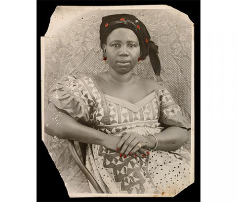 woman seated in front of patterned cloth, head wrapped in scarf, wearing printed dress, earrings, bracelet and ring, hands crossed over one another on her lap, details of cloth, earrings and fingernails hand tinted red