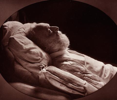profile view of an old man lying on a bed, propped up against pillows