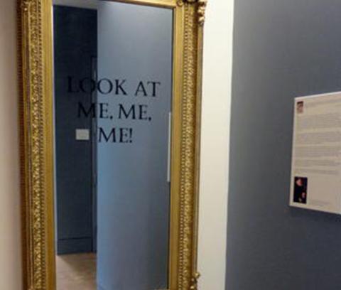 art gallery with mirror hanging on one of the walls. mirror has gold frame and black text on it that says, "LOOK AT ME, ME, ME!" 