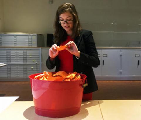woman with dark hair and glasses picks an orange slip of paper out of a red bucket that contains many orange slips of paper