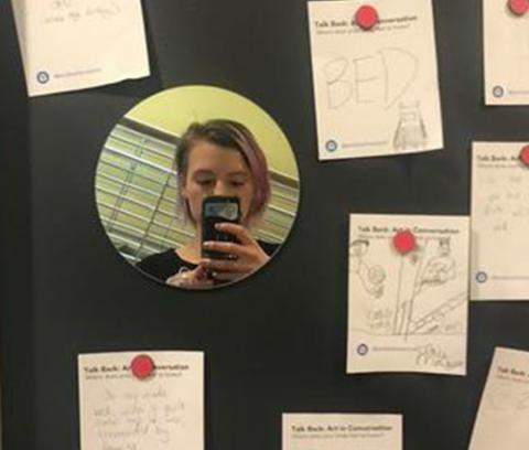 person holding a cell phone takes a picture of themselves in a mirror hanging on a black wall among pieces of paper tacked to the wall