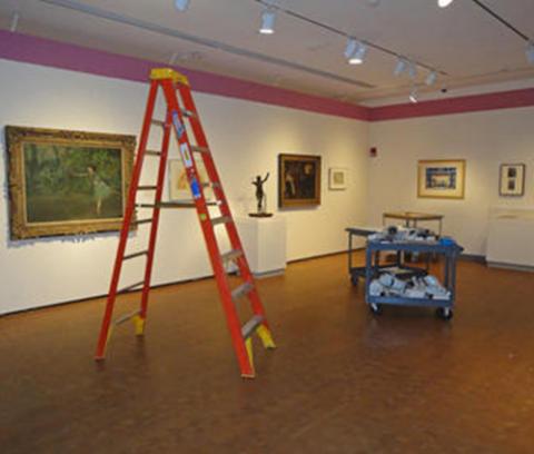 gallery with artworks displayed on the walls, orange ladder and blue cart in the middle of the gallery