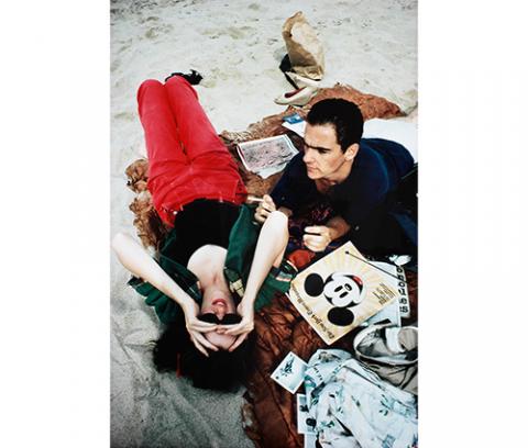 close-up of woman lying on her back on a rust colored quilt on a beach, she has dark hair, holds her sunglasses on her face with both hands, and wears red jeans, black leotard and green shirt, next to her is a man with short dark hair, lying on his stomach, holding a cigarette in his proper left hand, he has on a blue T-shirt and white shorts, scattered around them are shoes, newspaper sections, magazines, a paper bag, a bunched up coat, and brown slacks