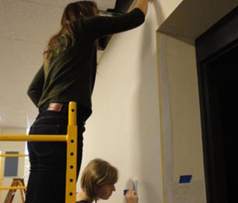 girl standing on a yellow ladder, drawing on a white wall. another girl stands below her, also drawing on the wall