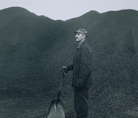 man in work clothes, gloves and hat standing facing proper right in huge pile of coal holding shovel in right hand