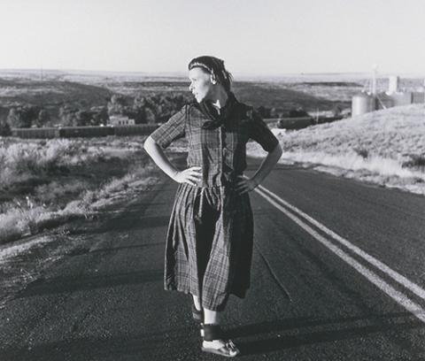 young woman with long plaid dress, sandals, ankle weights, hands on hips, head turned toward proper right standing in land of divided road with fields on either side and parts of buildings visible in low hills at distance