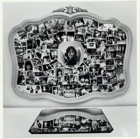 Mirror on a wall covered with photographs. Woman with her hands behind her head with a camera reflected in the center. Mirror on the surface below.