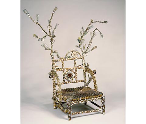 low seated chair made of partly painted branches with curving arms and extended branches at each corner and attached glass, bones, plastic, metal and dried creeper vine