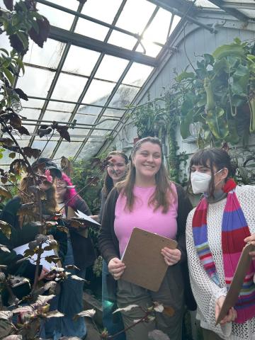 Three student museum educators smiling while holding clipboards and surrounded by plants in the Botanic Garden