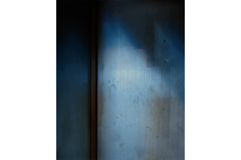 Photo of an interior room corner with the wall to the right perforated with small holes and covered in dark blue and brown shadows.