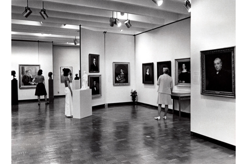 Black-and-white photograph of an art gallery with light walls and wood floor. Several painted portraits hang on the walls. Three figures stand throughout the space.