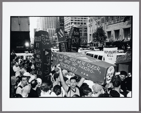 Photograph of protesters gathered on a crowded city street with cars in the background. Protest signs read, “Killed by Bush and Helms” and, “We’re here, we’re queer, we hate the fucking President!”
