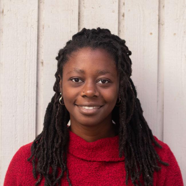 Should photo of a smiling young woman with long braids and a red sweater standing in front a a wall of white board and batton siding