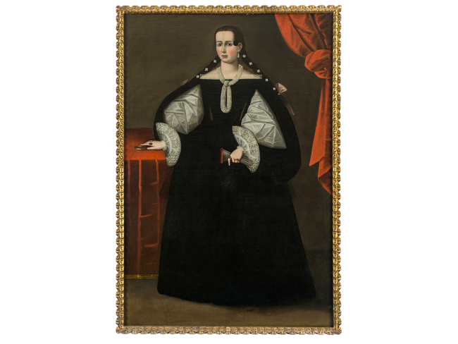 Person standing in elaborate black dress with white lace sleeves, pearl earring and necklace holding gloves in one hand