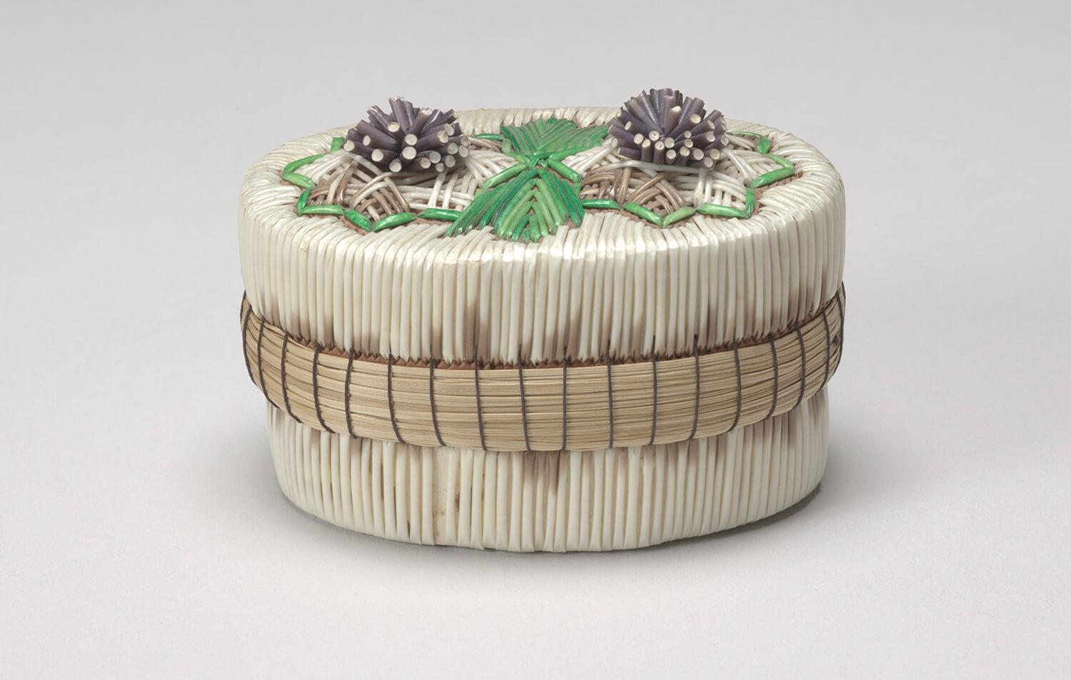 Woven basket with a lid, looks small with purple woven flowers and green leave on the top