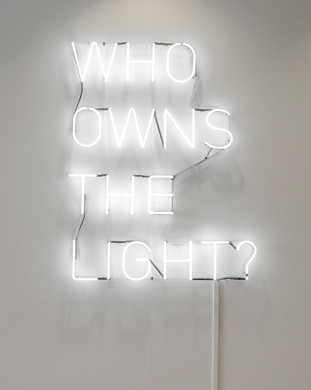 neon sign on a white wall with words stacks vertically that read, WHO OWNS THE LIGHT