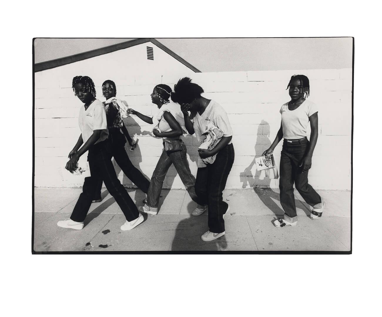black and white photo of 5 young girls dressed in black and white walking on a sidewalk with a white painted cement wall in background
