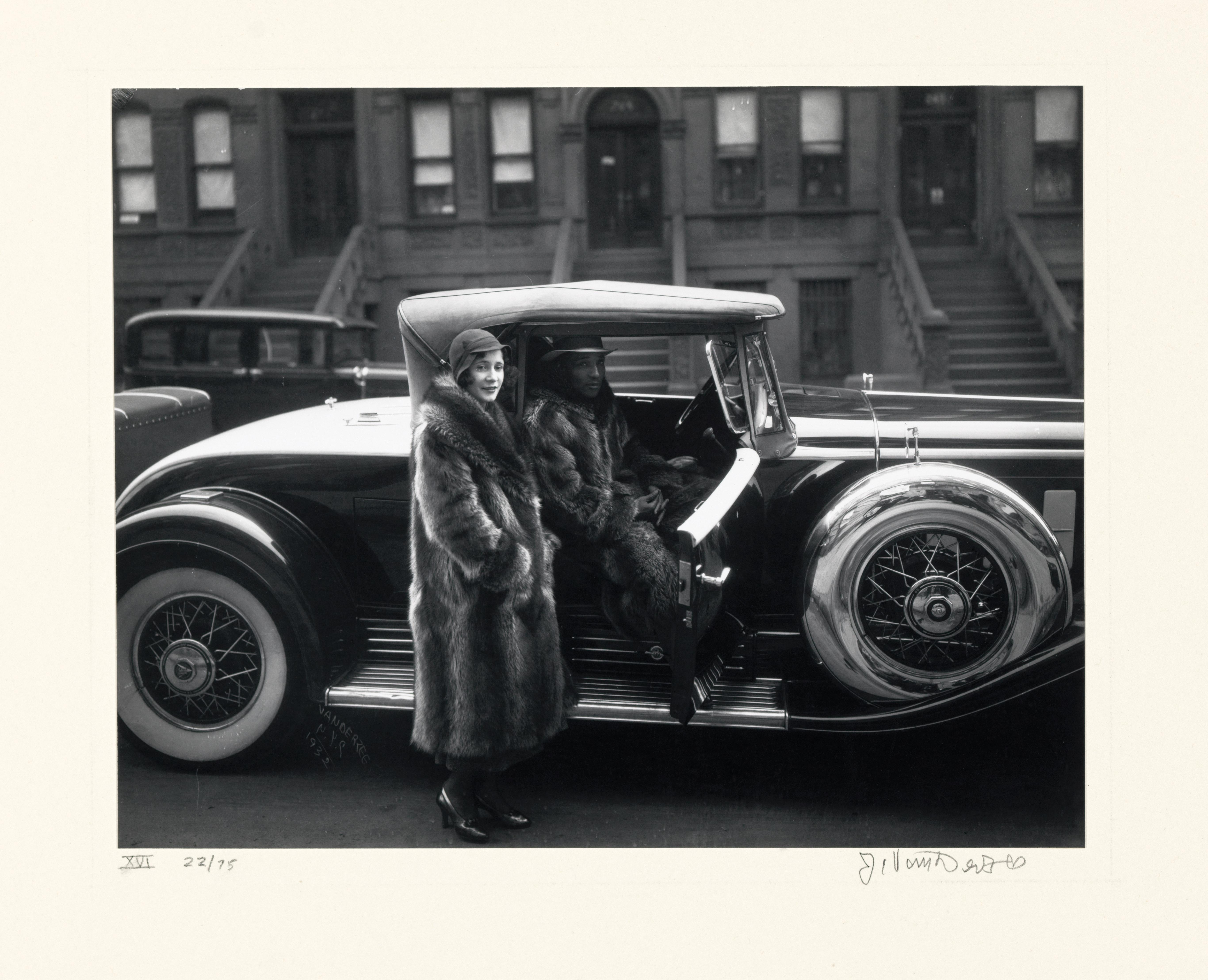 Black and white photo of a car from 1930's with woman standing next to the car in a fur coat and a man sitting inside the car