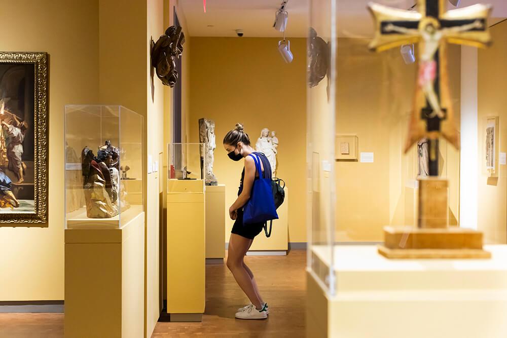 Young adult in shorts bending down to look closely at a gallery vitrine in a gallery with yellow walls