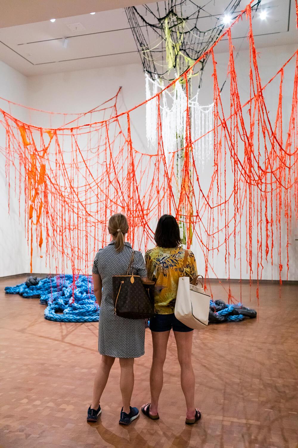 Backside of two people in gallery in frnot of an installation of red rope hanging from above and large blue rope draped on the floor