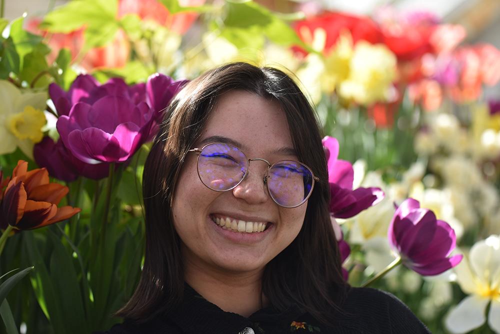 person with long brown hair and glasses smiling in front of tulips