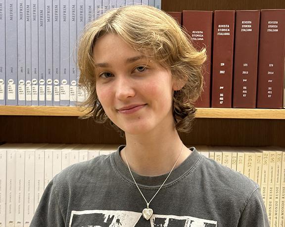 Person with short blond hair standing in front of a bookshelf