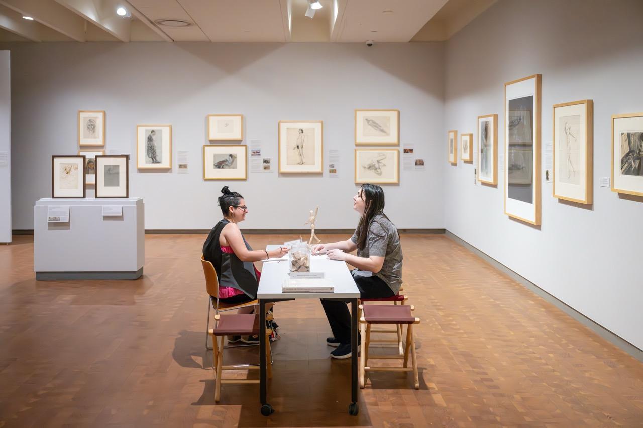 Two people sitting at a table drawing each other in the middle of a gallery with framed drawings on the walls