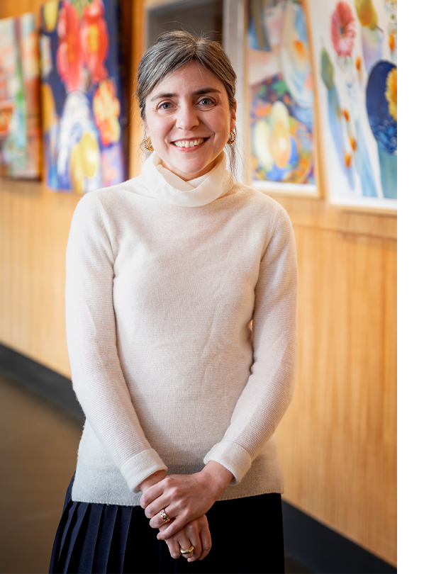 A person with white sweater with hands clasped in front of them standing in front of a wall of paintings
