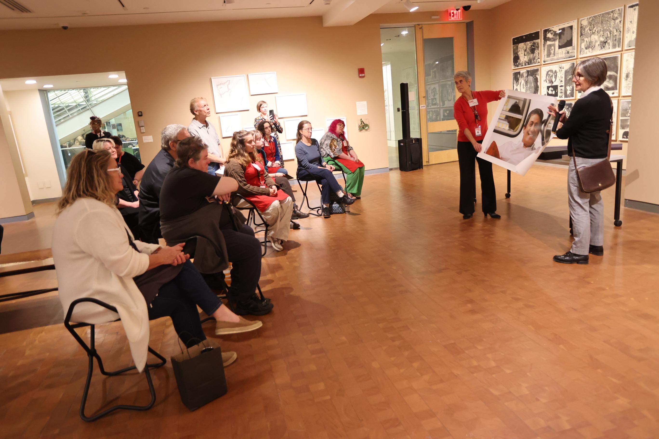 Gallery of people sitting and watching two people presenting a large 2-d reproduction of an artwork