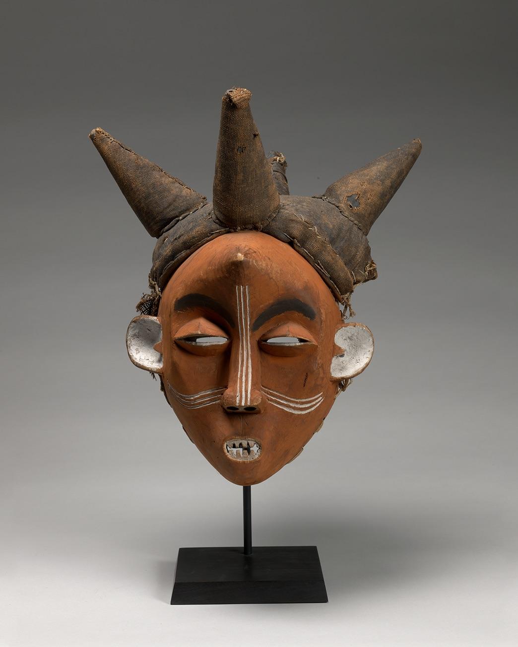 Handmade mask on a pedastal with ornage face aith three lines down nose and across cheeks and 4 point protruding from the hair and one point on the forehead