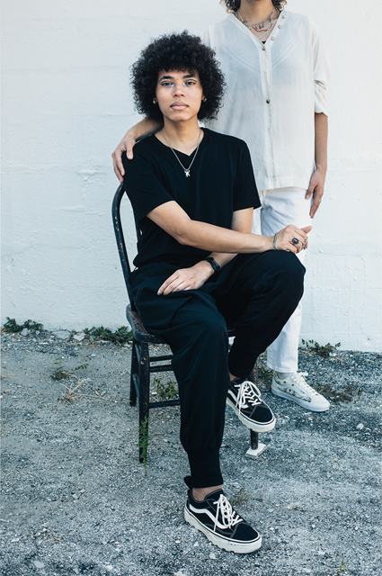 Photograph of two people, one with curley mediam length hair sitting in a chair dressed in back and the body of person behind them in white with their head showing in the photograph