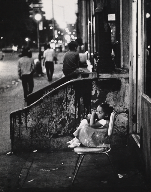 Black and white photo of a littered city street, with a two young children sitting next to and on worn concrete steps, while two adults walk down the street away from the viewer while street lights shine in the distance