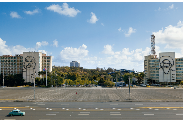 Streetscape with a baby blue old car in foreground with street in foreground, then an empty plaza and then buildings in background, two that have silhouettes of men on them