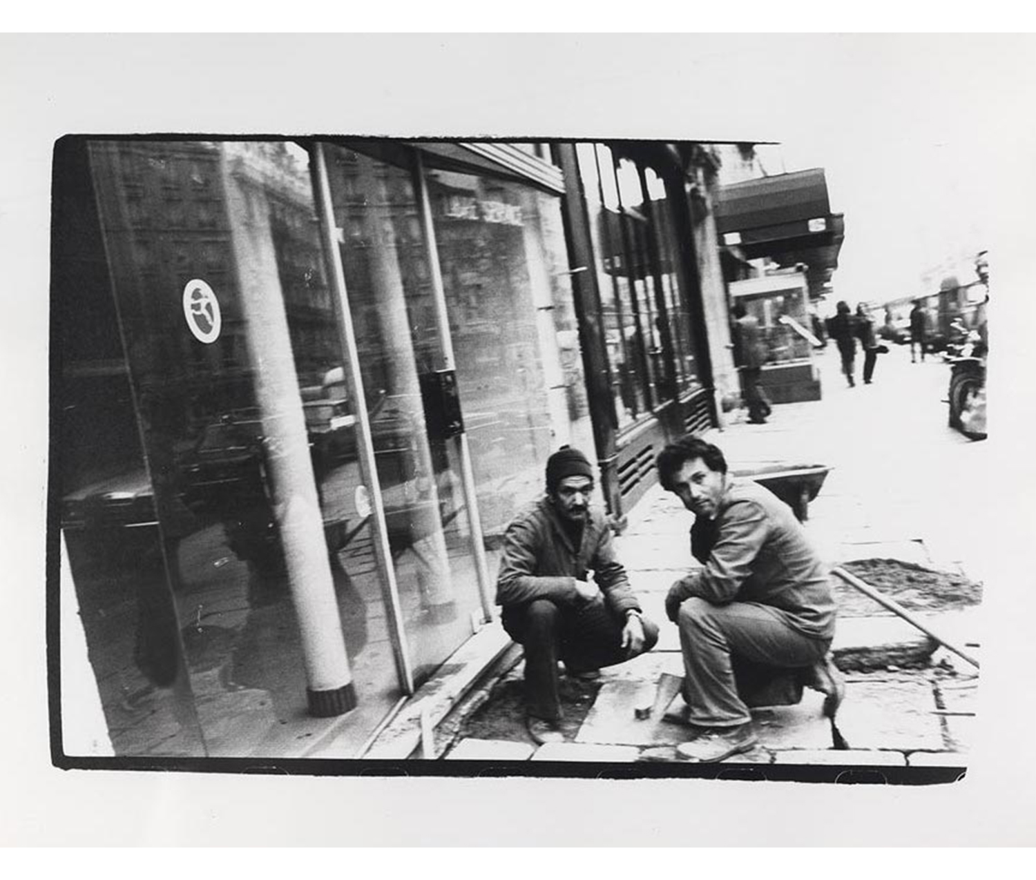 two men kneeling down next to each other on a busy city street, in front of a store