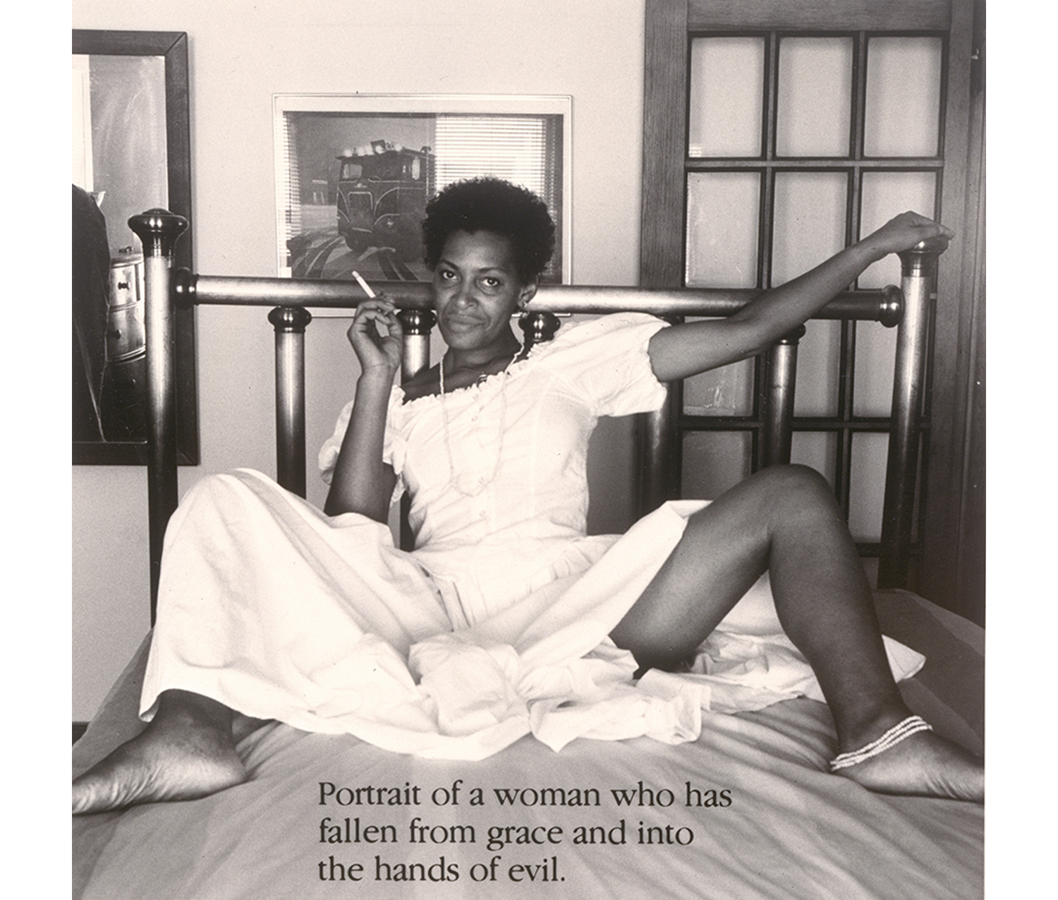 Black woman seated on bed with her legs spread, wearing a white dress and smoking a cigarette; gaze is focused at the camera