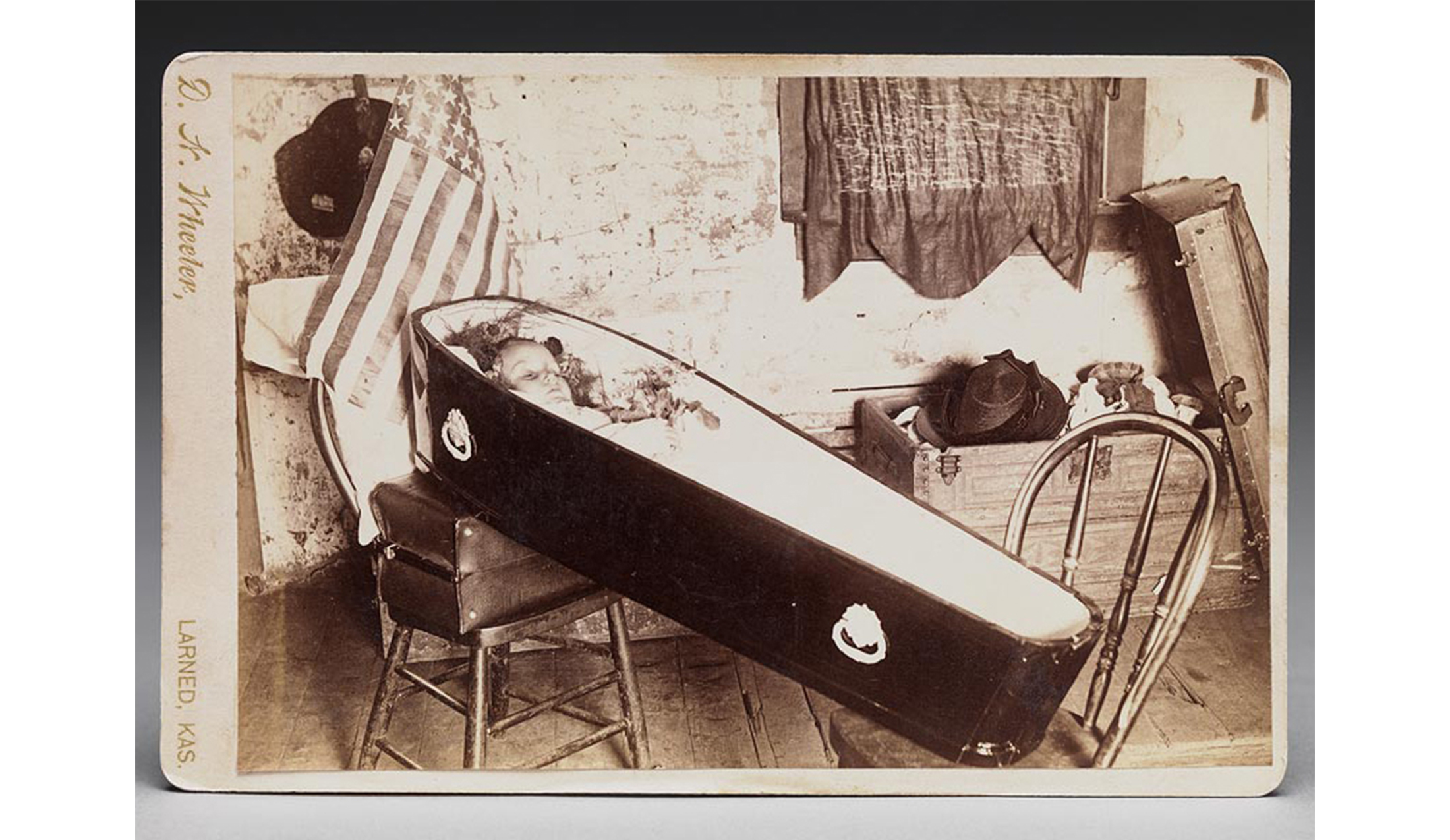 small black coffin holding a deceased dead child set on two chairs with American flag behind it and open trunk filled with children's clothing nearby, floor is unfinished wood, wall is heavily peeling whitewash, small window has translucent dark curtain over it