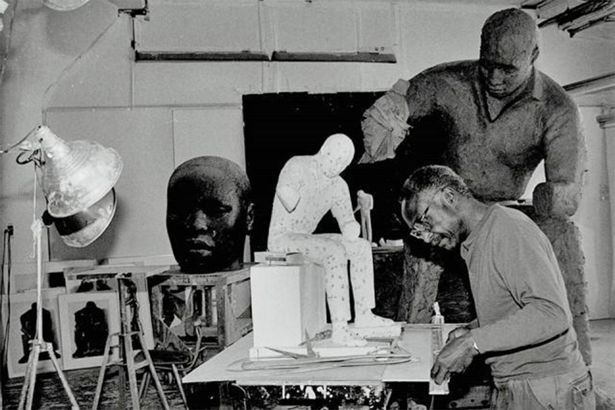 "black man leaning over desk measuring base of a sculpture with other sculptures are scattered through his studio"