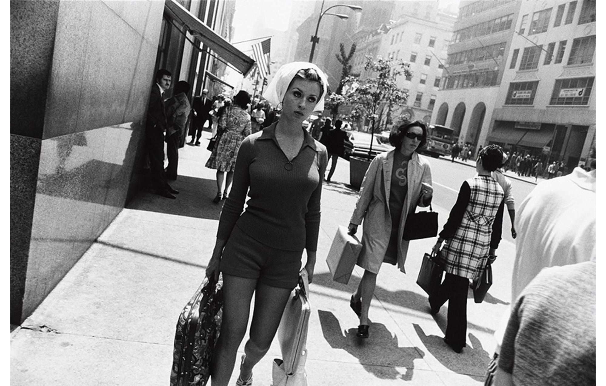 city sidewalk with pedestrians, young woman in front center, hair under scarf wearing shorts and knit top walks carrying paisley suitcase in one hand and two other bags in the other