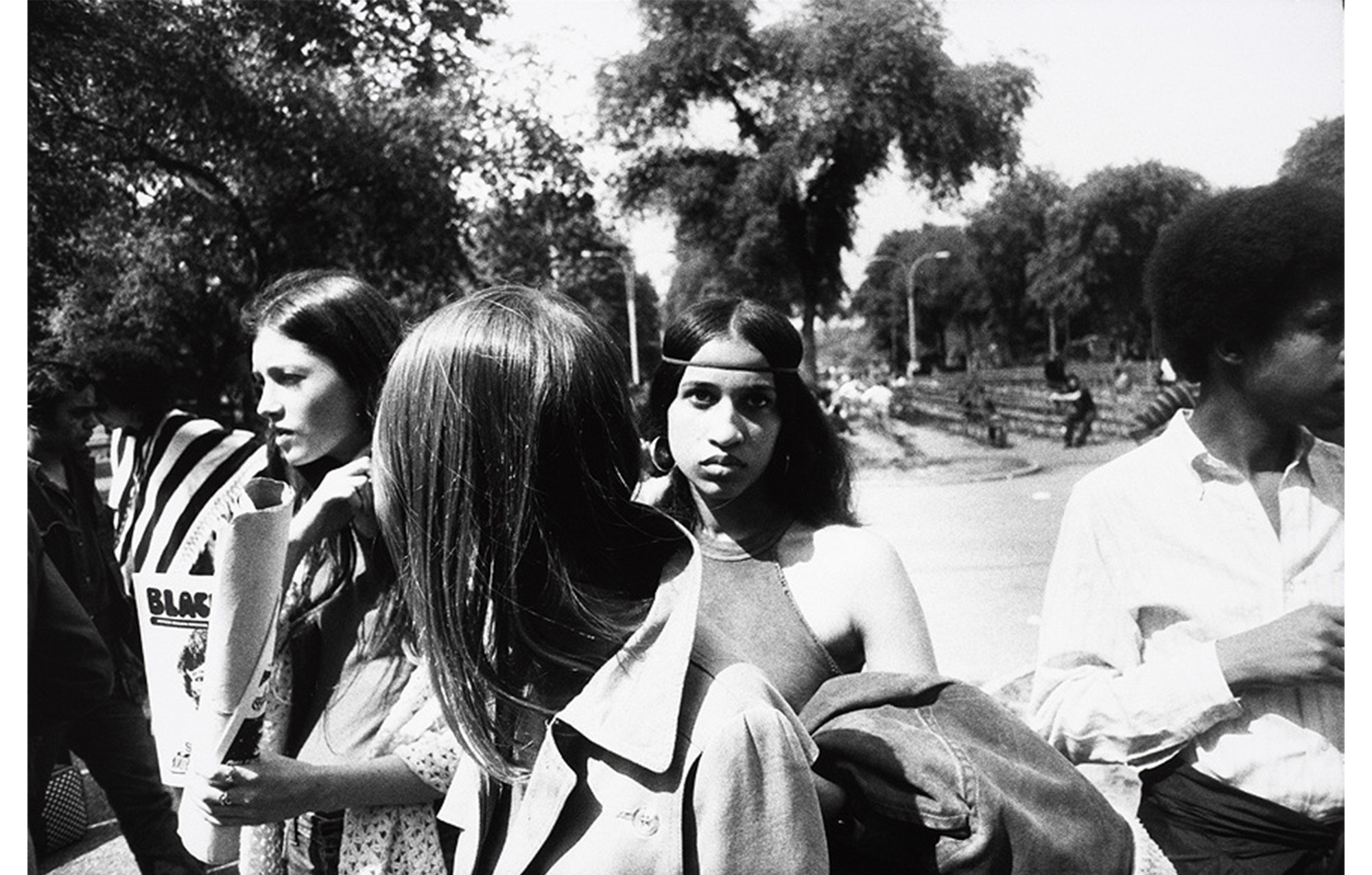 walkway in a park, close-up of group of young people, black woman in center with hair parted in middle and thin headband looking directly into the camera
