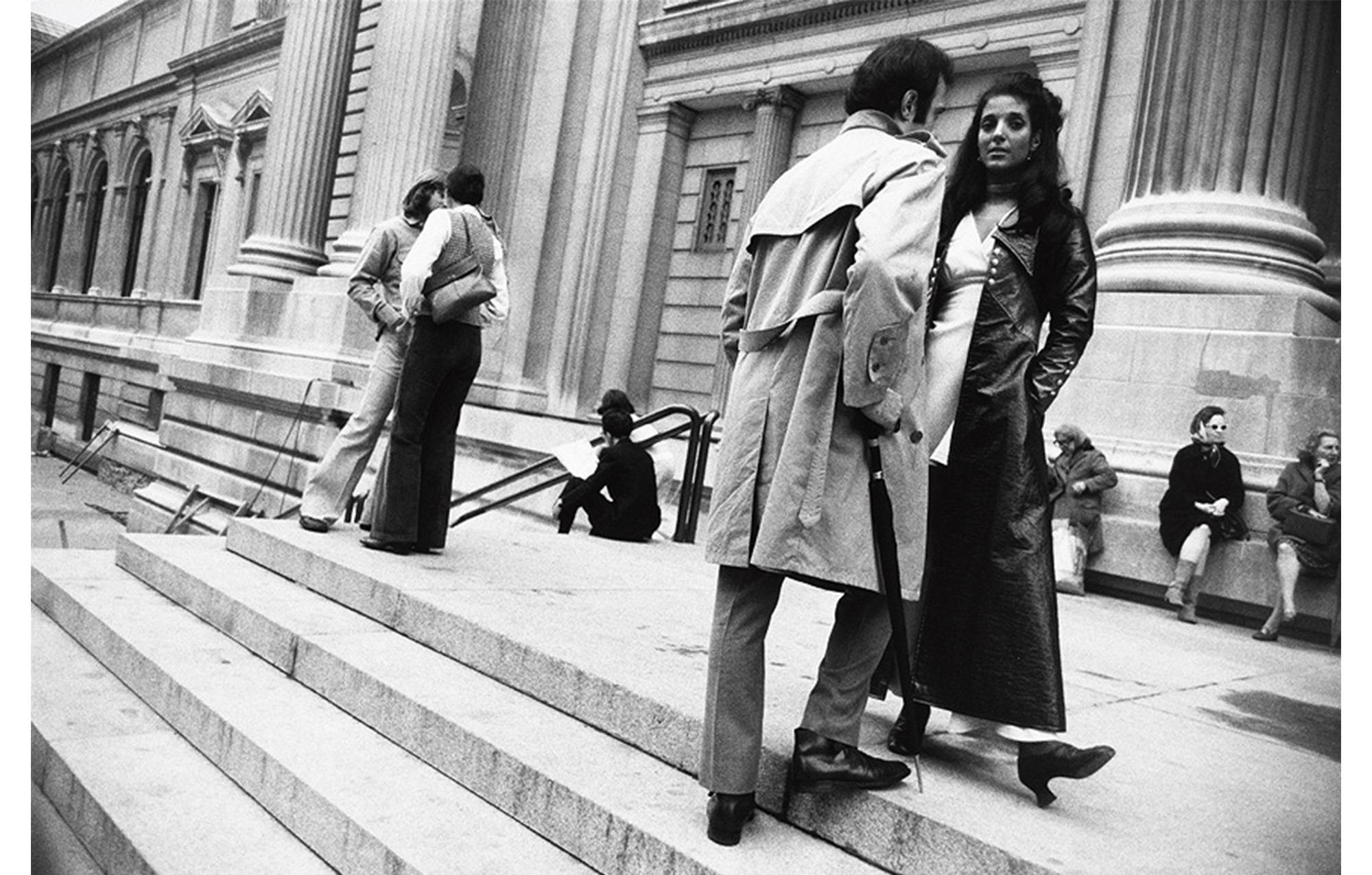 steps of the Metropolitan museum, two couples stand close together talking, front couple in coats, three older women sit near the column at back right and two people sit on steps in distance