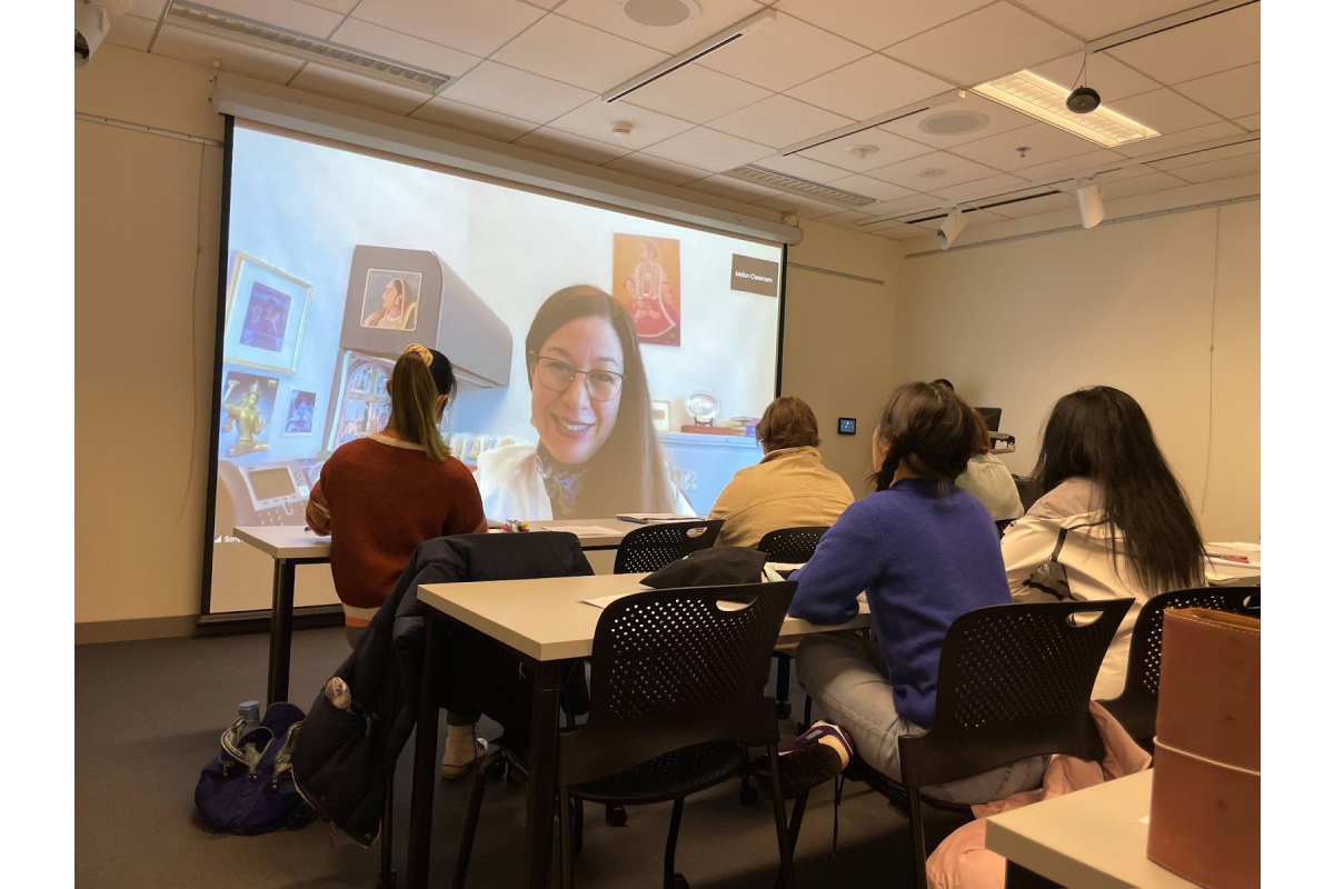 "students sitting at tables looking at a projection of a zoom meeting"